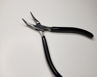Puede incluir: Unas tenazas de punta fina con mordazas curvas. Las tenazas tienen un cuerpo de metal plateado y mangos negros brillantes. La herramienta está diseñada para trabajos de precisión, con un enfoque en agarrar y doblar objetos pequeños.