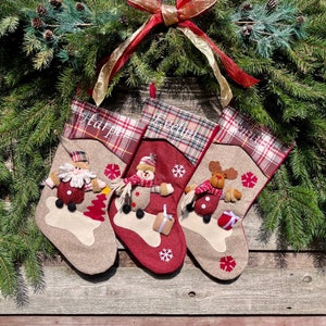 May include: Three red and brown Christmas stockings with personalized names "Harper", "Evelyn", and "Sawyer". Each stocking features a different festive character: Santa Claus, a snowman, and a reindeer.
