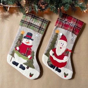 May include: Two personalized Christmas stockings with a snowman and Santa Claus design. The stocking on the left has the name "Hazel" embroidered on it and the stocking on the right has the name "Carter" embroidered on it.