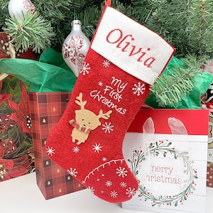 May include: Red and white Christmas stocking with the name Olivia embroidered on the cuff. The stocking features a reindeer with a scarf and the text "My First Christmas".
