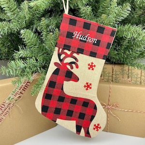 May include: Red and black buffalo plaid Christmas stocking with a reindeer design and the name "Hudson" embroidered on the cuff.