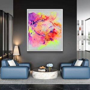 May include: Abstract painting in a white frame with vibrant colors of pink, purple, yellow, and orange. The painting is hanging on a dark gray wall above a coffee table and two blue armchairs.