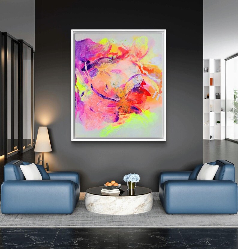 May include: Abstract painting in a white frame with vibrant colors of pink, purple, yellow, and orange. The painting is hanging on a dark gray wall above a coffee table and two blue armchairs.