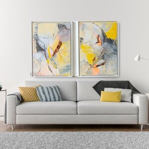 May include: Two abstract paintings in white frames, with yellow, gray, and orange brushstrokes, hanging above a white sofa with yellow and blue throw pillows.