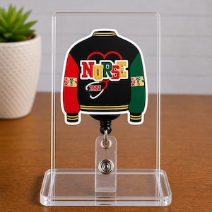 May include: A black, red, and green jacket-shaped badge reel with the word "NURSE" in yellow and white. The jacket design includes the text "BLACK NURSE MAGIC" and a stethoscope. The reel is displayed on a clear acrylic stand.