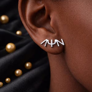 May include: Silver-toned ear climber earring with a minimalist design. The earring is shaped like a stylized set of eyelashes. The earring is worn on a person's ear, with a black background and gold-colored beads.