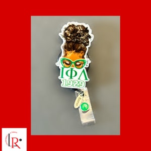 May include: A black retractable badge holder with a green and white design featuring a woman with glasses and the text "IΦΛ 1929". The holder has a green charm with the word "Soror" on it.