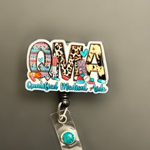 May include: A colorful badge reel with the letters "QMA" and the text "Qualified Medical Aide". The letters are decorated with medical symbols and patterns. The reel has a turquoise blue retractable cord and a turquoise blue button.