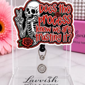 May include: A badge reel with a skull graphic giving a peace sign, surrounded by red roses. The text reads "Does the process know we are trusting it?" The reel has a clear plastic holder and a decorative, rhinestone-studded button.