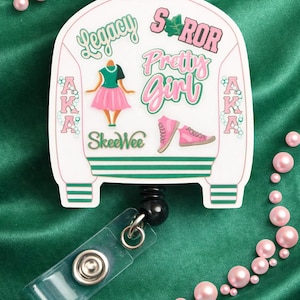May include: A white and green badge reel with a jacket design. The jacket has the words "Legacy," "Pretty Girl," and "SkeeWee" in pink and green. The badge reel also features a pink and green illustration of a woman in a dress and a pair of sneakers. The background is green.
