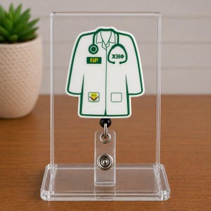 May include: A white and green medical badge reel featuring a doctor's coat design. The badge reel has the letters "NP" and "XHΦ" on the coat. The badge reel is displayed in a clear acrylic stand.