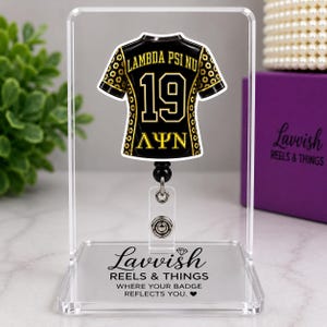 May include: A black and gold jersey-shaped badge reel featuring the text LAMBDA PSI NU and the number 19. The design includes Greek letters and a floral pattern on the sleeves. It is mounted on a clear acrylic stand with the text Lavvish REELS & THINGS.