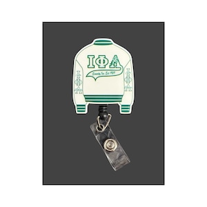 May include: A white and green badge reel featuring a jacket design. The jacket has green stripes and the Greek letters IΦA. The text "Serenity, Inc. Est. 1989" is also visible. The reel has a clear plastic strap.