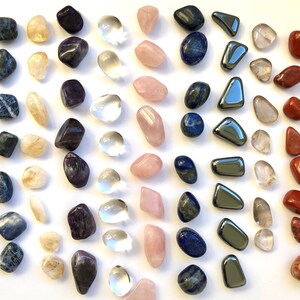 May include: Assortment of tumbled stones in various colours, including blue, purple, clear, pink, red, and grey. The stones are arranged in rows on a white background.