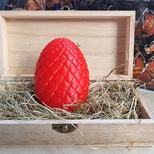 Op de afbeelding: Een rode drakenei-replica in een houten doos met stro. Het ei heeft een getextureerd, schubbig oppervlak.