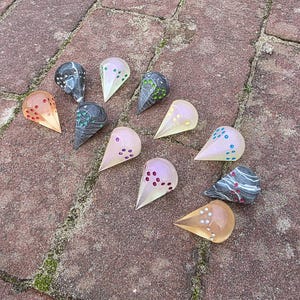 Op de afbeelding: Een verzameling druppelvormige glaskunstwerken in verschillende kleuren, waaronder oranje, roze en grijs, met decoratieve stippenpatronen. De stukken zijn gerangschikt op een bakstenen oppervlak.