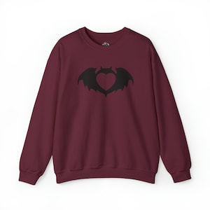 Puede incluir: Una sudadera color burdeos con un diseño de silueta de murciélago negro. El murciélago tiene una forma de corazón recortada en sus alas.