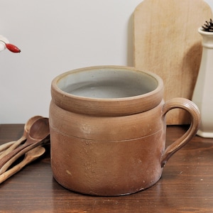 以下が含まれることがあります： 釉薬がかけられた内側と取っ手のある茶色の陶器の壺。壺は木製の表面の上に置かれています。