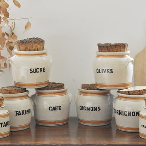 Può includere: Otto barattoli di ceramica bianca con coperchi di sughero e fasce arancioni. I barattoli sono etichettati con parole francesi: "Mostarda", "Farina", "Caffè", "Cipolle", "Cetrioli sottaceto", "Vaniglia", "Zucchero" e "Olive".