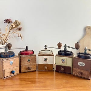 Puede incluir: Una colección de cinco molinillos de café Peugeot de madera vintage con tapas y cajones de varios colores. Cada molinillo tiene una manivela y un cajón para recoger el café molido. Los molinillos se exhiben sobre una superficie de madera.