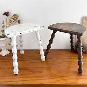 May include: Two small, wooden step stools. One is painted white with distressed details, the other is a dark brown. Both have a half-moon shaped seat and turned legs. Decorative items are in the background.