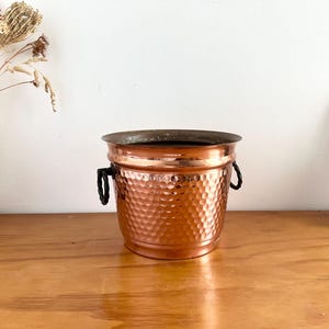 May include: A copper bucket with a hammered finish and two black handles. The bucket has a wide rim and is on a wooden surface. The bucket is empty and appears vintage.