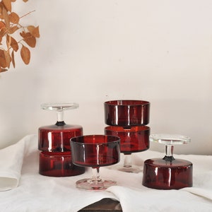May include: Five red glass dessert bowls with clear glass stems. The bowls are stacked in pairs and arranged on a white linen tablecloth.