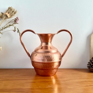 May include: A hammered copper vase with two handles. The vase is a light copper colour and has a textured surface.