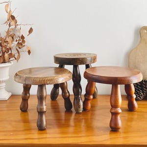 Puede incluir: Tres taburetes de madera con patas torneadas. Los taburetes son de color marrón y tienen un acabado de madera natural. Están dispuestos en fila sobre una superficie de madera.