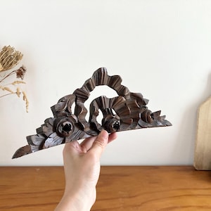 Könnte beinhalten: Ein dunkelbrauner architektonischer Giebel aus Holz mit aufwendigen, handgeschnitzten Blumenmotiven und geschwungenen Blattmustern. Das Dekorationsobjekt hat eine dreieckige Form mit symmetrischem Design und zeigt detaillierte Holzmaserungen.