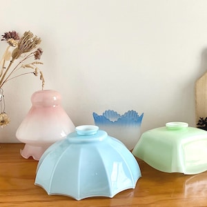 Op de afbeelding: Een verzameling vintage glazen lampenkappen in verschillende pastelkleuren. De lampenkappen omvatten een lichtblauwe parapluvormige kap, een lichtgroene vierkante kap en een roze ombre kap. Een witte vaas met gedroogde bloemen en een houten snijplank staan op de achtergrond.