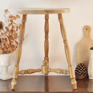 Può includere: Uno sgabello in legno con un sedile rotondo e tre gambe. Lo sgabello è realizzato in legno marrone chiaro e ha un design semplice.