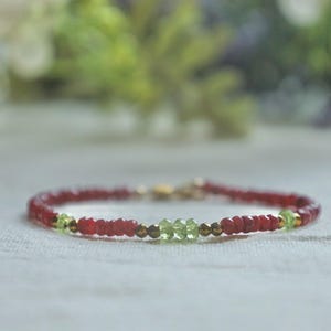 May include: A delicate bracelet featuring small, round, red beads interspersed with green and gold-colored beads. The bracelet is secured with a gold clasp. The background is blurred, with hints of green and white.
