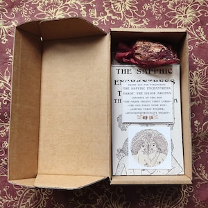 以下が含まれることがあります： 茶色の厚紙の外側を持つ開いた箱には、「The Sapphic Enchantress Tarot: The Major Arcana」というタイトルのタロットデッキが収められています。箱には、白いステッカーに「Sapphic Tarot Sticker」と書かれた小さな赤いポーチも含まれています。