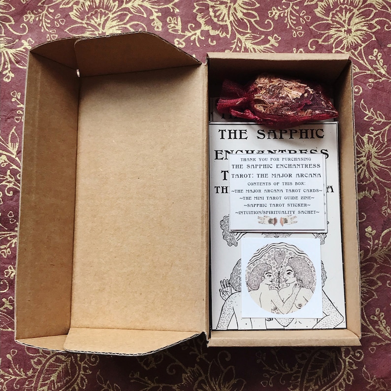 以下が含まれることがあります： 茶色の厚紙の外側を持つ開いた箱には、「The Sapphic Enchantress Tarot: The Major Arcana」というタイトルのタロットデッキが収められています。箱には、白いステッカーに「Sapphic Tarot Sticker」と書かれた小さな赤いポーチも含まれています。