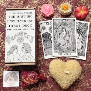 以下が含まれることがあります： 「The Sapphic Enchantress Tarot Deck」というタイトルと「The Major Arcana」というサブタイトルが書かれたタロットデッキの箱。箱が開いており、2人の人物が抱き合っている様子が描かれている。3枚のタロットカードが見え、1枚は「The World」というタイトル、もう1枚は「Lovers」というタイトルが書かれている。画像には、ハート型のろうそくなど、小さなろうそくがいくつかある。