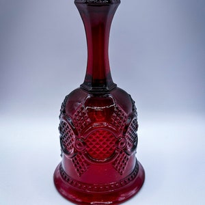 Puede incluir: Una campana de cristal rojo rubí con un diseño texturizado y estampado. La campana presenta una base acampanada, un cuerpo detallado y un cuello largo que conduce a una parte superior redondeada y texturizada. La campana es un artículo decorativo.
