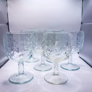 May include: Set of six clear, textured wine glasses. Each glass features a round bowl with a dimpled pattern and a stem. The glasses are arranged on a white surface, with a soft, diffused light.