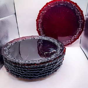 May include: A stack of round, deep red glass plates featuring a scalloped edge and a decorative embossed border pattern. One additional matching red glass plate stands upright behind the stack, displaying the same intricate rim design.