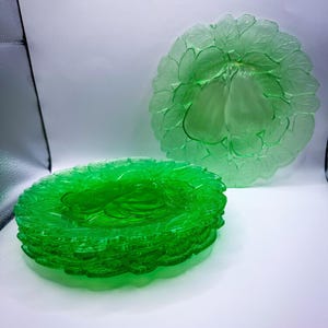 May include: A stack of bright green glass plates with a leaf design. A single plate with a pear design is visible in the background. The plates are made of a translucent green glass.