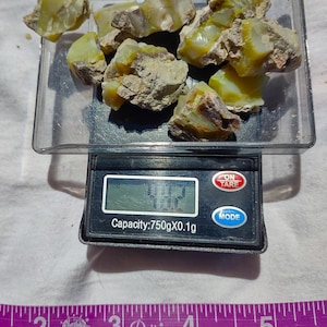 May include: A digital scale with a capacity of 750g x 0.1g, displaying a weight of 4.27 grams. The scale is on a white surface with a ruler and a pile of green and yellow rough opal stones in a clear plastic container.
