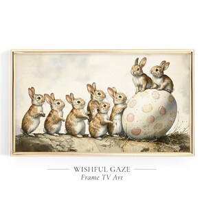 Peut inclure: Une peinture à l'aquarelle d'un groupe de lapins bruns regardant un gros œuf blanc avec des taches roses et jaunes. Les lapins sont disposés en ligne, le plus grand lapin étant debout sur l'œuf. La peinture est encadrée d'un cadre doré et porte le texte "Wishful Gaze" et "Frame TV Art" sous l'image.
