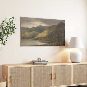 May include: A landscape painting in a frame shows a tranquil mountain scene with a lake and forests. The artwork, in muted tones, is above a wooden cabinet with woven doors, alongside a white lamp and decorative plants.