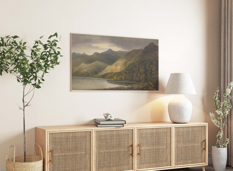 May include: A landscape painting in a frame shows a tranquil mountain scene with a lake and forests. The artwork, in muted tones, is above a wooden cabinet with woven doors, alongside a white lamp and decorative plants.