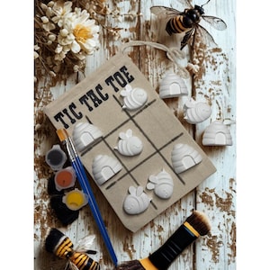 May include: A tan fabric tic tac toe game board with black grid lines and the text TIC TAC TOE. The board features white bee and beehive shaped game pieces. Included are a blue paintbrush, small paint pots, and two decorative bee figures.