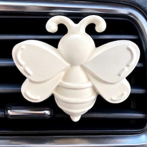 May include: A cream-colored bee-shaped car air freshener clip featuring a smooth, matte finish with embossed wing details and a segmented body, securely attached to the horizontal slats of a vehicle's dashboard air conditioning vent.