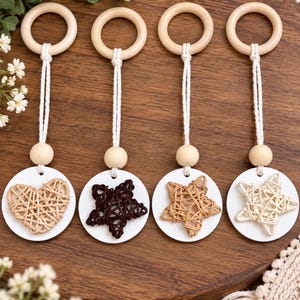 May include: Four hanging ornaments featuring a light wood ring at the top, a white cord, a small round wooden bead, and a white circular base. The ornaments display a woven heart, a dark brown woven star, and two light woven stars.