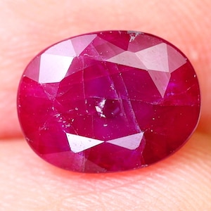 Może przedstawiać: Owalny, ciemnoczerwony rubin o fasetowanym szlifie. Rubin ma kilka widocznych inkluzji.