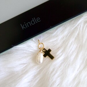 May include: A gold-colored charm with a cross and a pearl, attached to a Kindle e-reader. The e-reader is black with the word "kindle" printed on it. The background is white, fluffy fabric.