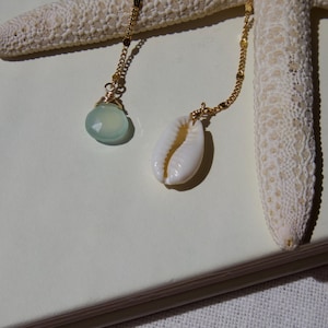 May include: Two gold necklaces with ocean-themed pendants rest on a white surface next to a starfish. One necklace features a light blue gemstone pendant, while the other has a white seashell pendant.
